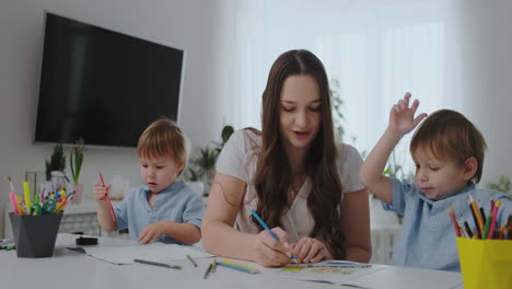 A-young-mother-with-two-children-sitting-at-a-white-table-draws-colored-pencils-on-paper-helping-to-do-homework